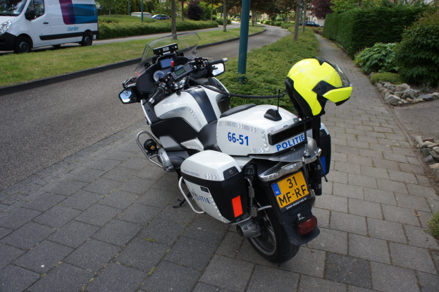 Polizeimotorrad am Straßenrand mit einem Helm darauf, umgeben von Bäumen, Strommasten, Gebäuden, Vegetation, anderen Fahrzeugen.