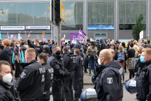 Eine große Gruppe von Menschen steht vor einem Gebäude, einige halten Schilder und tragen Helme, mit einem Mast mit einem Schild im Vordergrund und einem Baum im Hintergrund, die zu protestieren scheinen.