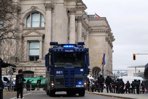 Gruppe von Polizeibeamten vor einem großen Gebäude mit architektonischen Details, Fahrzeugen auf der Straße, einer Person mit einer Kamera links und Hintergrundelementen wie Bäumen, Verkehrssignalen, Fahnenmasten und einem klaren Himmel.