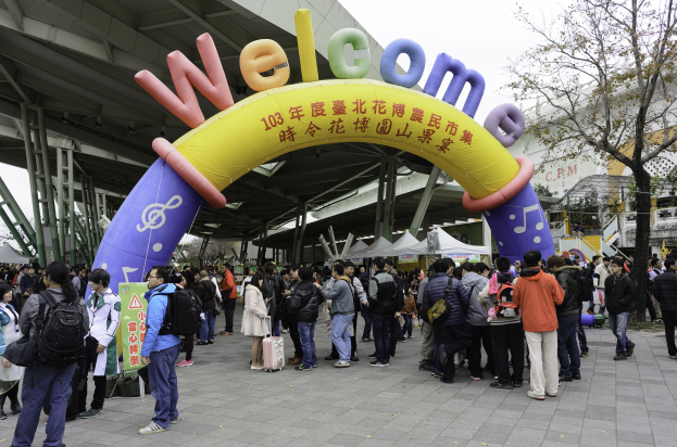 Gruppe von Menschen vor einem Willkommensbogen auf einem Outdoor-Event mit Zelten, Bäumen und Gebäuden unter einem klaren blauen Himmel.