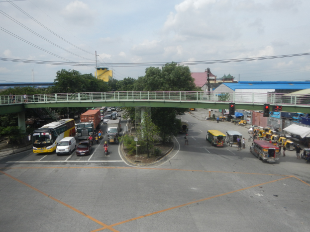 Eine belebte Stadtstraße mit Autos, Bussen, Fußgängern auf einem Gehweg, eine Brücke mit Geländern, Verkehrsampeln, Bäume, Gebäude, Strommasten mit Drähten und eine bewölkte Himmel.