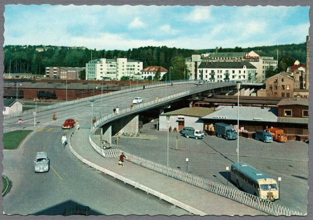Altes Schwarz-Weiß-Foto einer Stadtstraße mit Fahrzeugen, Fußgängern auf einer Brücke, Gebäuden, Bäumen, Laternenmasten und einem bewölkten Himmel.
