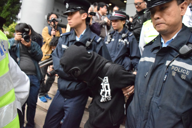 Eine Gruppe von Polizisten steht vor einer Menge, einige halten Kameras, mit einem Gebäude mit Säulen und Bäumen im Hintergrund.