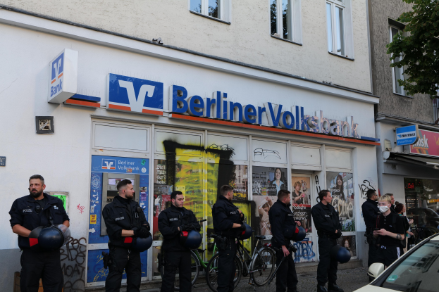 Gruppe von Polizeibeamten vor einer Berliner Volksbank stehend, einige halten Helme in den Händen, mit einem Auto auf der rechten Seite und einem Baum auf der linken Seite, Gebäude mit Fenstern und Texttafeln im Hintergrund.
