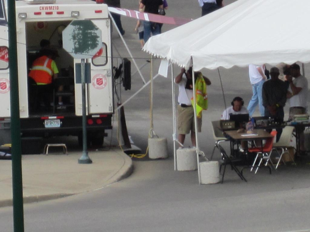 Ein Rettungswagen steht auf der linken Seite einer Straße, während auf der rechten Seite ein Zelt eine Gruppe von Menschen schützt, die in der Nähe eines Tisches und eines Stuhls stehen.