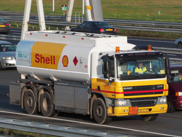 Shell-Tanklastwagen fährt auf einer Autobahn mit anderen Fahrzeugen, die sichtbare Insassen enthalten, und fährt an Straßeninfrastruktur wie Geländern, Pfählen, Schildern und Grasflächen vorbei.