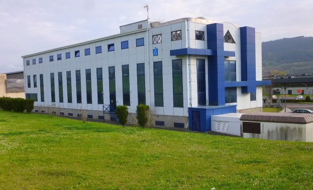 Großes weißes und blaues Gebäude auf einem grünen Feld mit Bäumen, Fahrzeugen, Polen, Lampen, Schildern, Bergen und einem klaren blauen Himmel im Hintergrund.