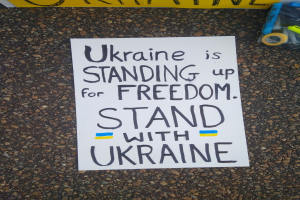 Ein weißer Papierbogen mit den Worten "Ukraine steht für Freiheit, stellt Euch mit Ukraine" liegt vor einer gelben Fahne auf dem Boden, rechts daneben eine Plastikabdeckung mit verschiedenen Gegenständen.