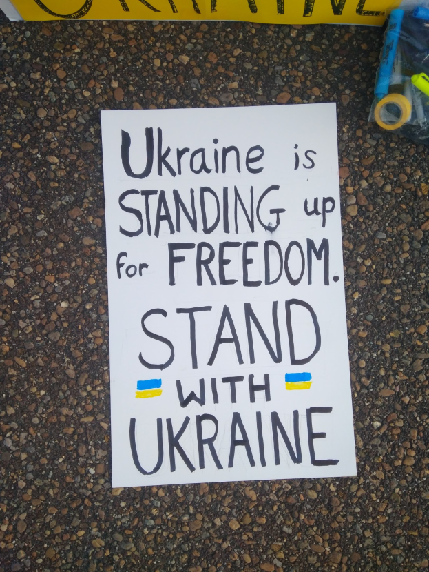 Ein weißer Papierbogen mit den Worten "Ukraine steht für Freiheit, stellt Euch mit Ukraine" liegt vor einer gelben Fahne auf dem Boden, rechts daneben eine Plastikabdeckung mit verschiedenen Gegenständen.
