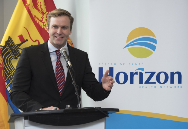 Ein Mann mit einem schwarzen Blazer, weißem Hemd und roter Krawatte steht an einem Podium mit einem Mikrofon und einem Banner im Hintergrund und lächelt.
