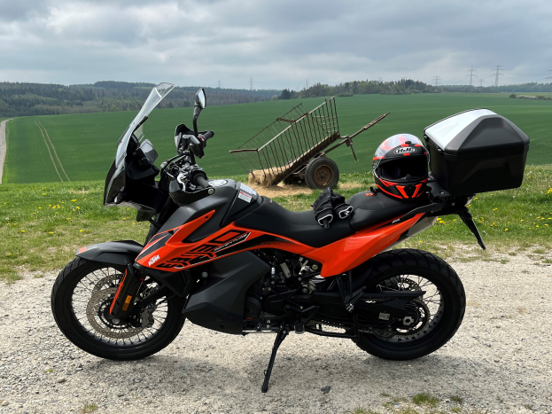 Ein KTM 1290 Super Adventure R Motorrad auf einem Schotterweg mit einem Helm und einem Gepäckträger daneben, umgeben von grünem Gras, hohen Bäumen und Strommästen mit Drähten unter einem bewölkten Himmel.