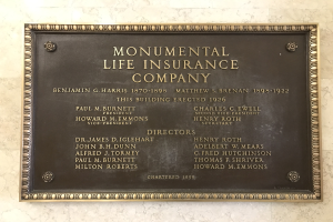 Plakette an einem Gebäude mit der Aufschrift "Monumentale Lebensversicherung" in fetter Schrift, angebracht auf einem dunklen Material mit einem dekorativen Rahmen.