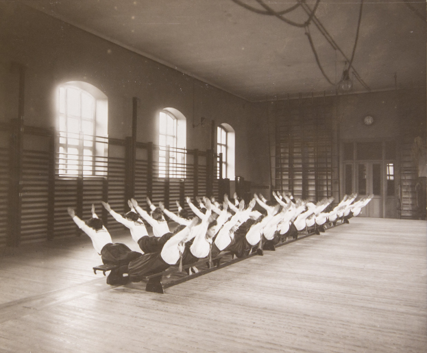 Gruppe von Menschen bei Pilates-Übungen in einem Fitnessstudio mit Fenstern und einer Uhr an der Wand.
