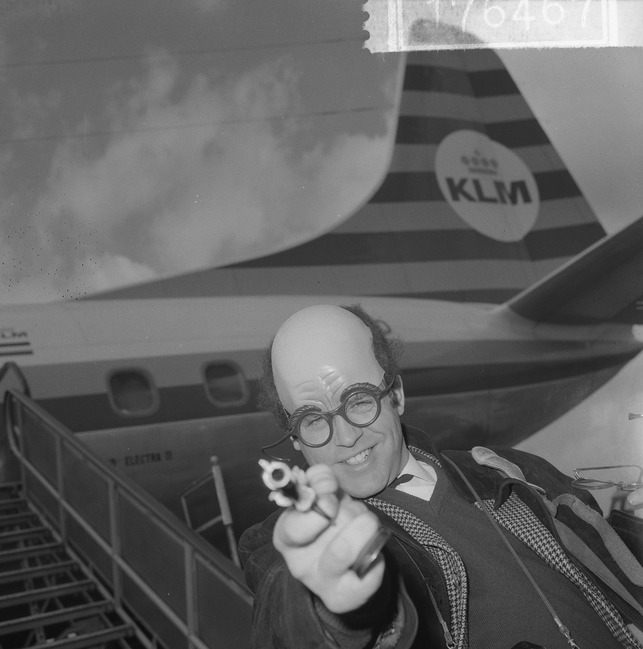 Schwarzes und weißes Bild eines Mannes in Anzug und Krawatte, der eine Waffe vor einem startenden Flugzeug hält.