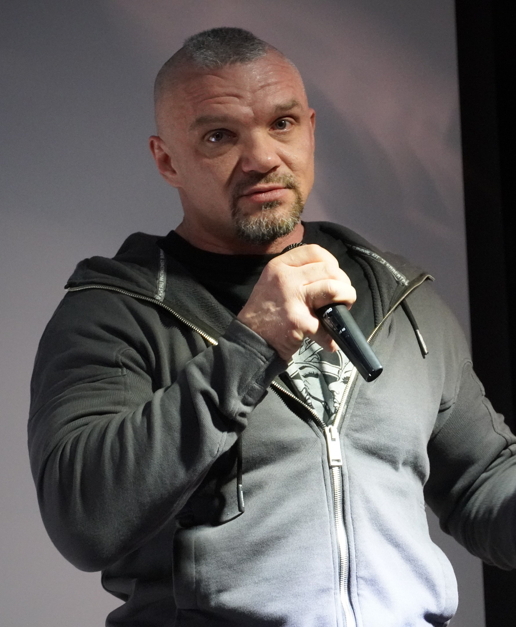 Ein Mann in einer schwarzen Kapuzenjacke und einem schwarzen Anzug mit einem weißen Hemd hält ein Mikrofon und steht selbstbewusst vor einer Wand, er scheint ein Komiker mit zurückgekämmtem Haar zu sein.