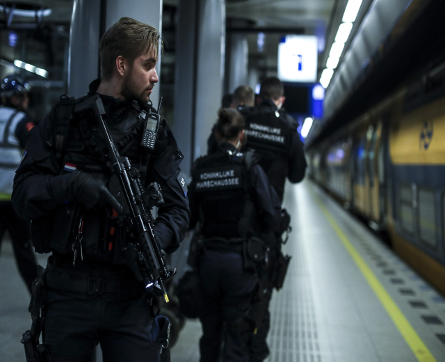 Polizeibeamte in schwarzen Uniformen, einige mit Waffen, stehen auf einem Bahnsteig mit beleuchteten Lichtern und Säulen im Hintergrund.