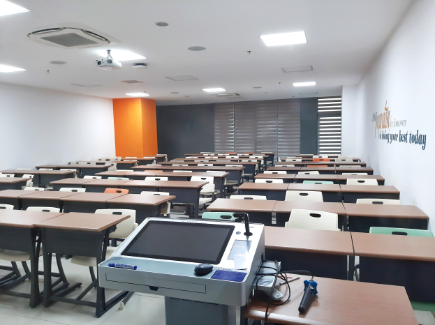 Ein Klassenzimmer mit Tischen und Stühlen in Reihen, ein deckenmontierter Projektor und Gegenstände auf Tischen, mit Text an der rechten Wand, der darauf hinweist, dass es sich um eine Privatschule in Singapur handelt.