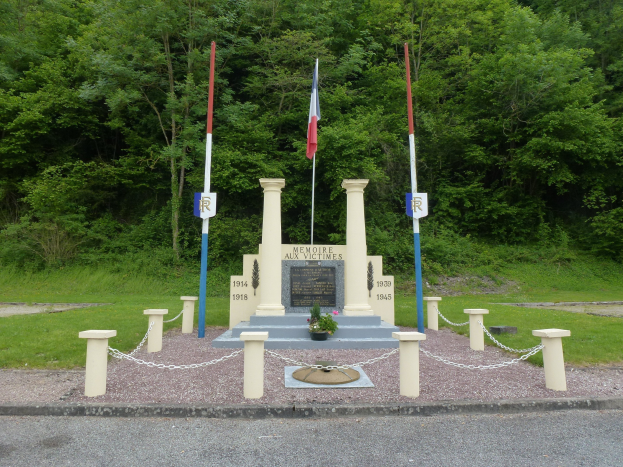 Gedenkstein mit eingravierter Inschrift, umgeben von Kettenpfählen und einem Blumentopf, mit drei Fahnen davor, in einem grasbewachsenen Park mit Bäumen im Hintergrund.