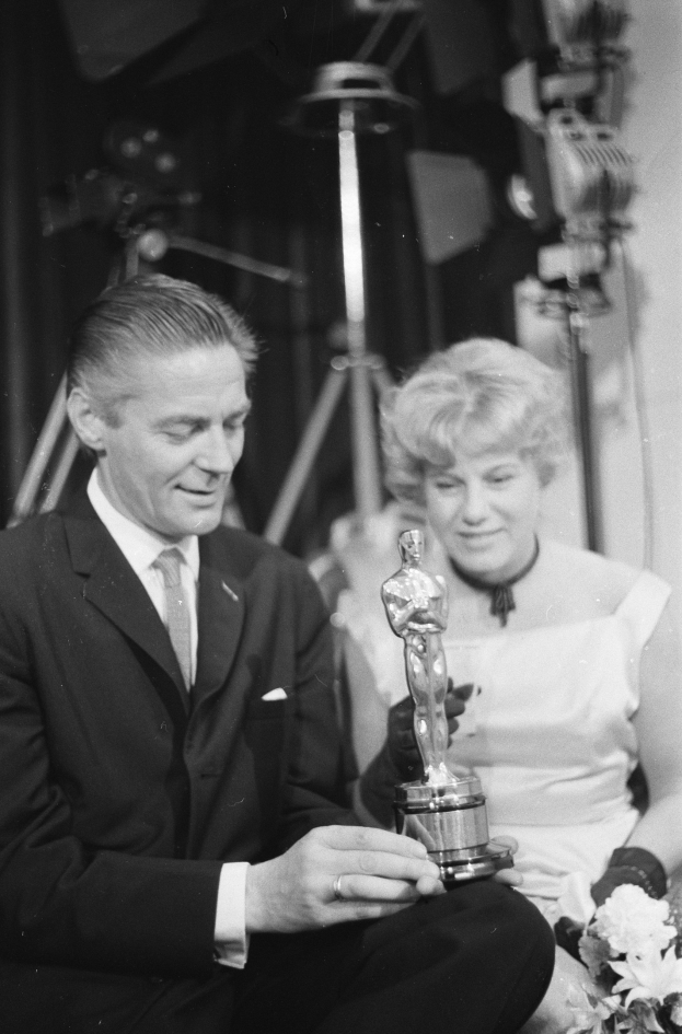 Ein Mann in einem schwarzen Blazer, weißem Hemd und Krawatte hält eine Oscar-Statue in der Hand und sitzt neben einer Frau, die einen Blumenstrauß hält, beide in eleganter Kleidung, mit Lichtern im Hintergrund, dargestellt in Schwarz-Weiß.