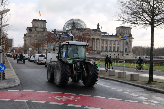 Ein Traktor fährt auf einer Straße in Berlin, Deutschland, vor dem Reichstag-Gebäude vorbei, das mit Flaggen geschm√√Ľckt ist, während Menschen zu Fuß gehen und Fahrr√°der fahren auf dem baumgesäumten Gehweg unter einem bewölkten Himmel.