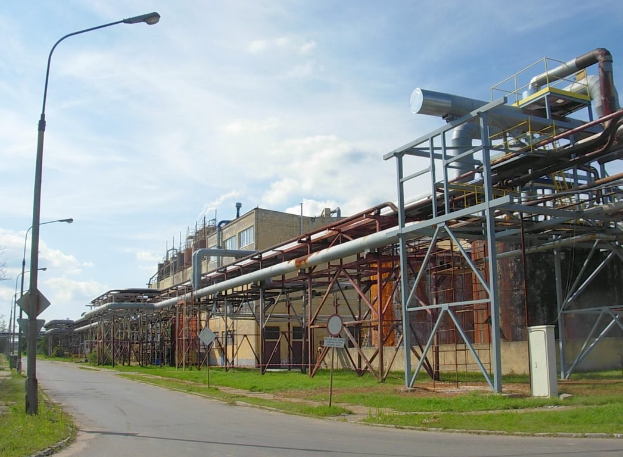Große Fabrik mit ausgedehnten Rohrleitungen, umgeben von Gebäuden, Straßeninfrastruktur, Grünflächen und einem bewölkten Himmel.