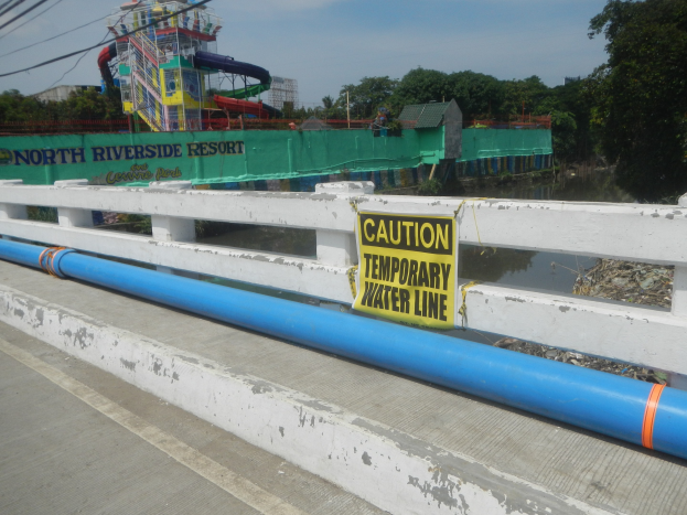Warnschild mit der Aufschrift "Vorsicht temporäre Wasserleitung" auf einer Brücke mit Geländer, Bäumen, Gebäuden, einer Wasserrutsche und einem bewölktem Himmel im Hintergrund.
