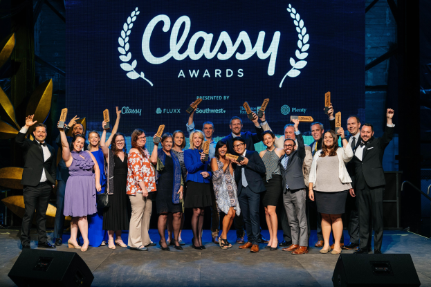 Gruppe von Menschen auf einer Bühne, die Auszeichnungen halten, mit Lautsprechern darunter und einem Bildschirm im Hintergrund mit der Aufschrift "Classy Awards 2019".