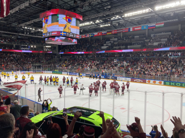 Hockey game in a large arena with spectators, rink fencing, banners, ceiling lights, a screen, and hanging flags, with a car visible in front of the crowd.