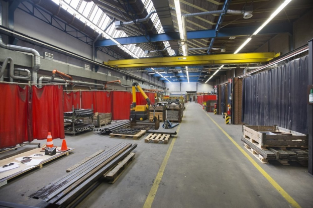 Großer Lagerraum mit Holzpaletten, Metallobjekten, Verkehrskegeln und Maschinenfahrzeug, mit roten Vorhängen, Rohren, Deckenleuchten und einer verhängten Wand.