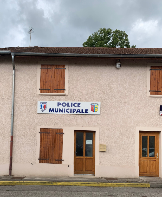 Gebäude der Polizei mit einem Schild, Fenstern, Türen, einer Namensplakette, einem Rohr, einem Baum und einem bewölkten Himmel im Hintergrund.