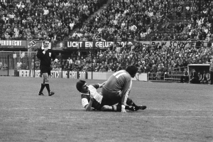 Ein Schwarz-Weiß-Foto von zwei Fußballspielern im Einsatz, einer stellt den anderen, mit Zuschauern und Stadion-Elementen im Hintergrund und einer Person, die ein Objekt auf der linken Seite hält.