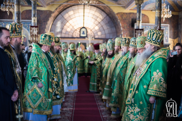 Priester in grünen und goldenen Gewändern stehen in einer Säulenkirche mit Kronleuchtern, während eine Frau rechts ein Mobiltelefon hält.