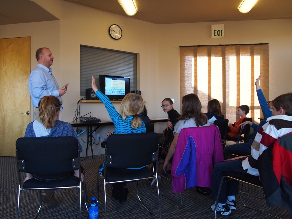 Ein Mann steht in einem Klassenzimmer und spricht zu einer Gruppe von Kindern, die auf Stühlen sitzen, mit einem Monitor, Lautsprechern, einer Uhr, einem Fenster und einer Tür im Hintergrund.