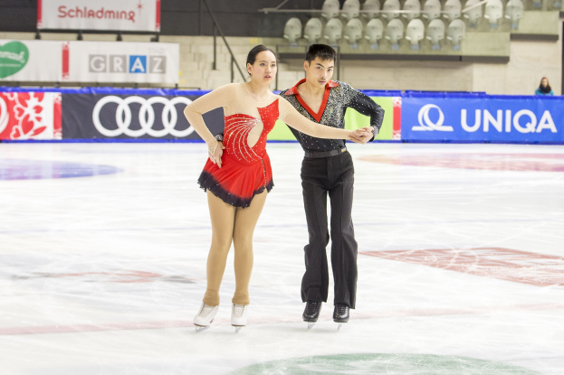 Zwei Eiskunstläufer, ein Mann und eine Frau, gleiten Hand in Hand über eine Eisbahn mit einer Menge und Absperrungen im Hintergrund.