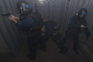 Eine Gruppe von Polizeibeamten in Einsatzausrüstung, einer hält eine Waffe in der Hand, steht neben einem Metallcontainer und einer Leiter in einem begrenzten Raum.