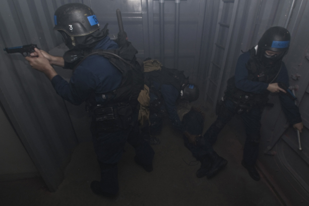 Eine Gruppe von Polizeibeamten in Einsatzausrüstung, einer hält eine Waffe in der Hand, steht neben einem Metallcontainer und einer Leiter in einem begrenzten Raum.
