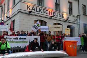 Eine Gruppe von Menschen mit Schildern und Transparenten vor einem Gebäude während einer Demonstration in Deutschland, mit zwei Personen auf einem Objekt im Vordergrund und einem Müllcontainer rechts.