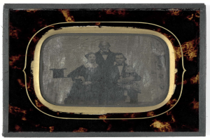 Schwarze-weiße Fotografie eines Mannes in Anzug und Krawatte und einer Frau in unbekannter Kleidung, die vor einem Fernseher stehen und ernste Gesichter haben.