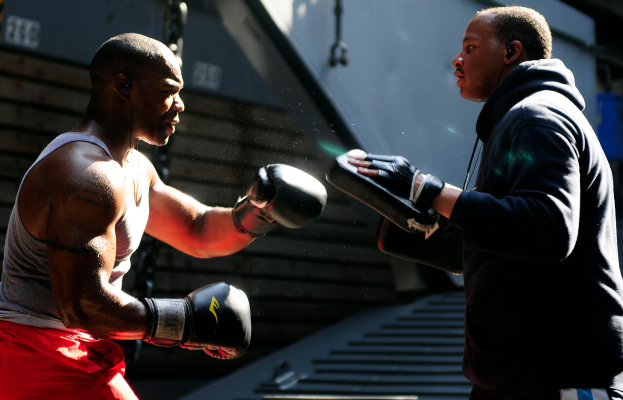 Zwei Männer in einem Boxring mit Handschuhen, die aufeinander einprügeln, mit einer Treppe im Hintergrund und einem blauen Objekt auf der rechten Seite.