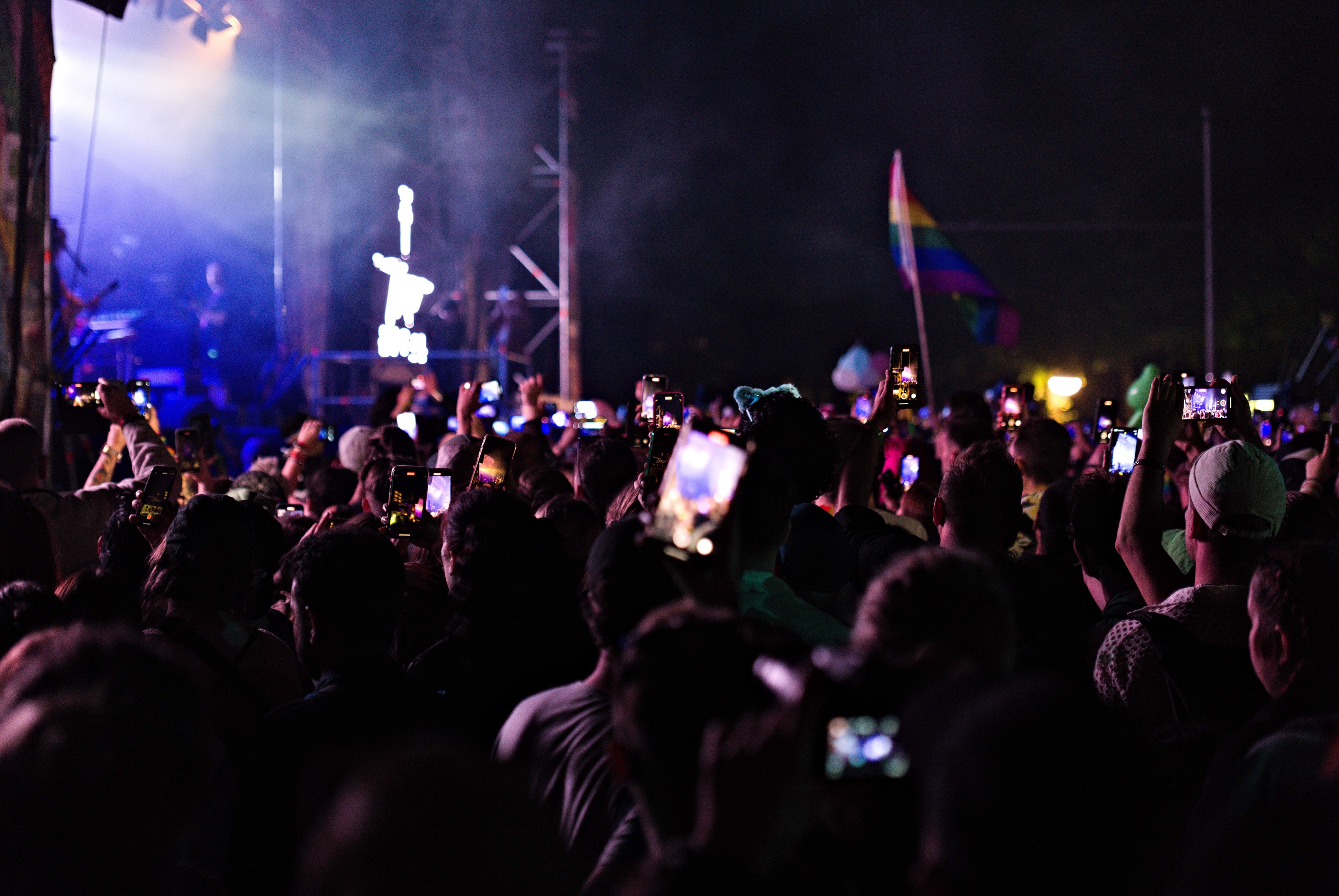 Menschenmenge auf einem Konzert mit hochgehaltenen Handys, beleuchtet von Lichtern, umgeben von Pfählen und einer Fahne, während Musiker auf der Bühne spielen.
