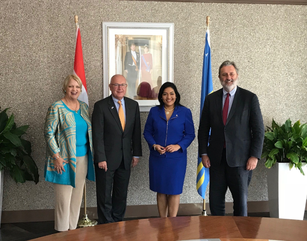 Gruppe von Menschen, die um einen Tisch mit Papieren stehen, umgeben von Fahnen, Topfpflanzen und einem Rahmenfoto an der Wand während einer Unterzeichnung.