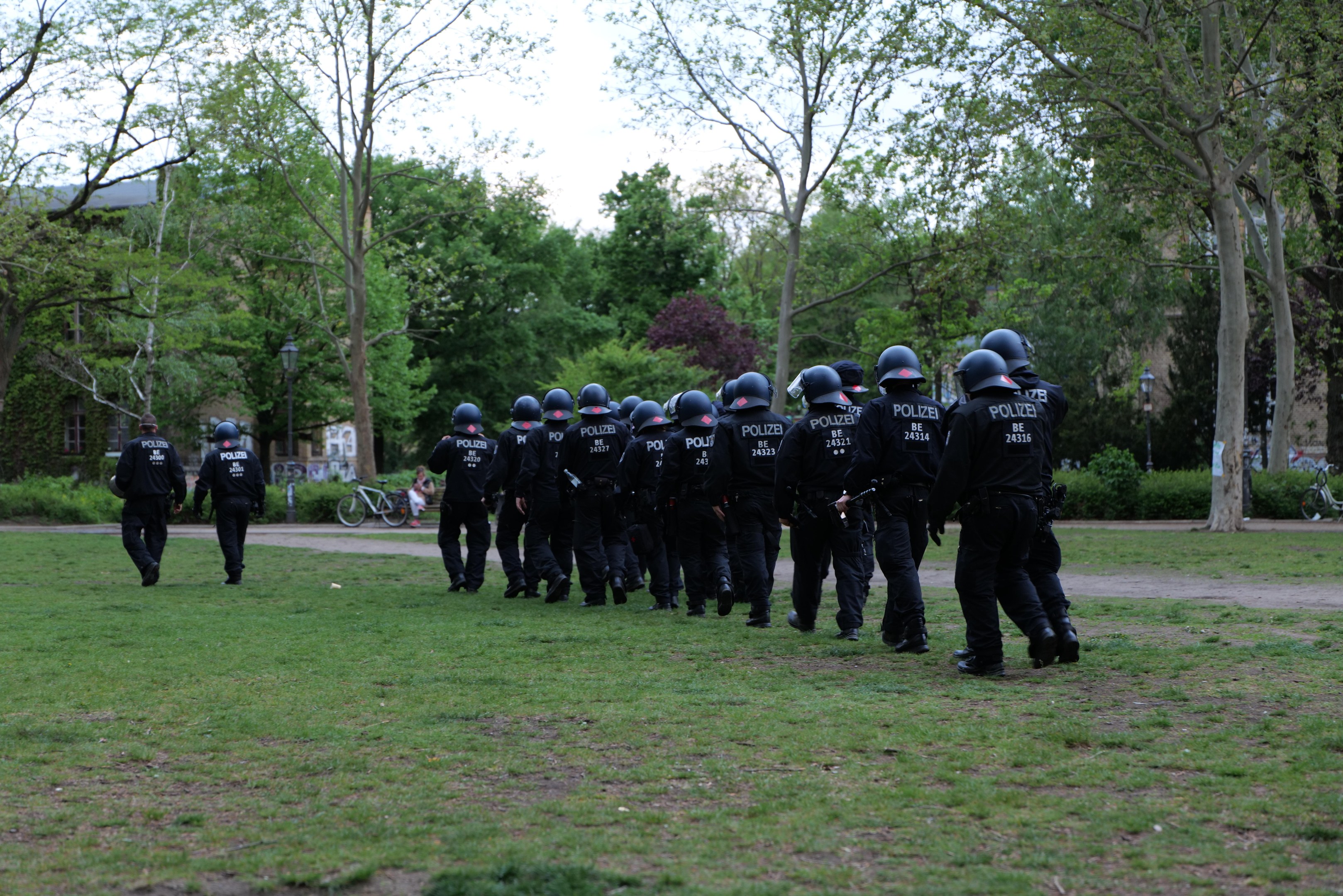 Eine Gruppe von Polizisten in schwarzen Uniformen und Helmen, die über ein grünes Feld gehen, mit Fahrrädern, Laternenmasten, Pflanzen, Bäumen, Gebäuden und einem klaren blauen Himmel im Hintergrund.