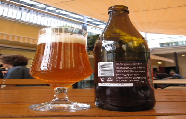 Ein Glas Wein und eine Weinflasche auf einem Tisch mit einer Gruppe von Menschen, einem Zelt, einem Gebäude und Lichtern im Hintergrund.