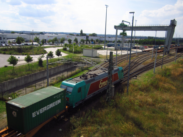 Güterzug fährt auf Schienen neben einem grünen Feld mit Strommasten, Kabeln, Gras, Bäumen, Gebäuden, Fahrzeugen und einem bewölkten Himmel.